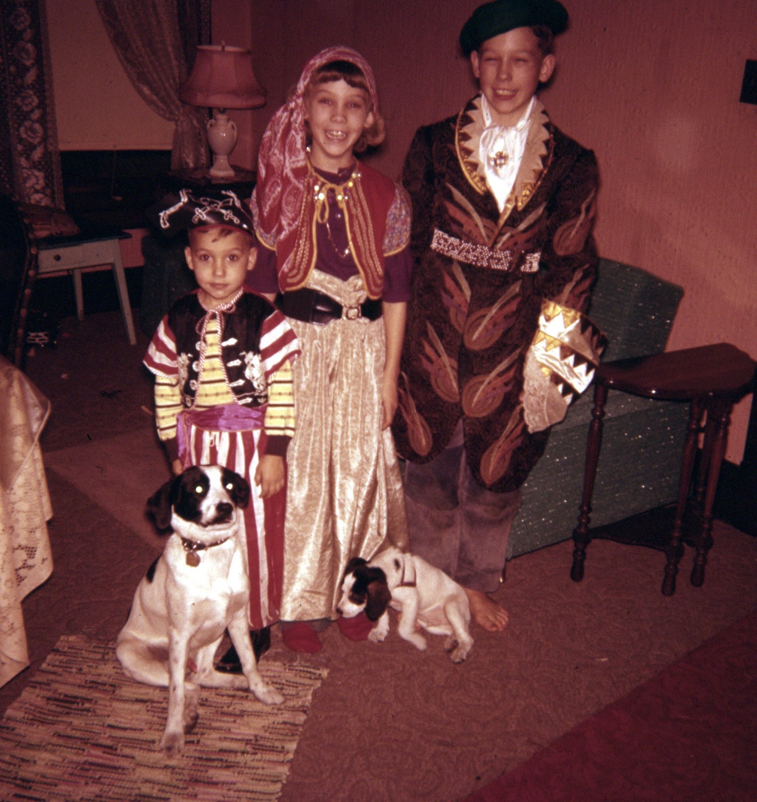 three siblings in 1958 pose in their self-created Halloween costumes in 1958. A 5 year-old boy is dressed in a pirate's hat, sash and striped pants; the girl, Linda, is dressed in the image of a gypsy, with bandana, flowing skirt, sash at waist, colorful vest; the oldest boy, 12, is dressed in a buccaneer's hat, an oversized brocaded jacket, shimmery shirt with lace cuffs; two spotted dogs, father & son, named Buttons and Bows, are at their feet; Bows is a puppy, about 8 weeks old.