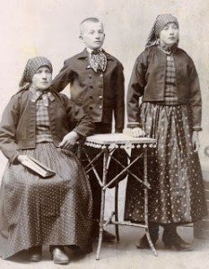 An early 1900 photo of the Gaertz family, ethnic Germans from Transylvania, around 1904. The widowed mother is seated with Bible in hand; her son, Josef Gaertz, is standing with hi right arm on his mom's shoulder. His sister is to his right. Both women are dress in traditional clothing of the era: long skirts to their ankles; short, waist-length jackets, and scarves covering the heads, tied at the nape of the neck.
